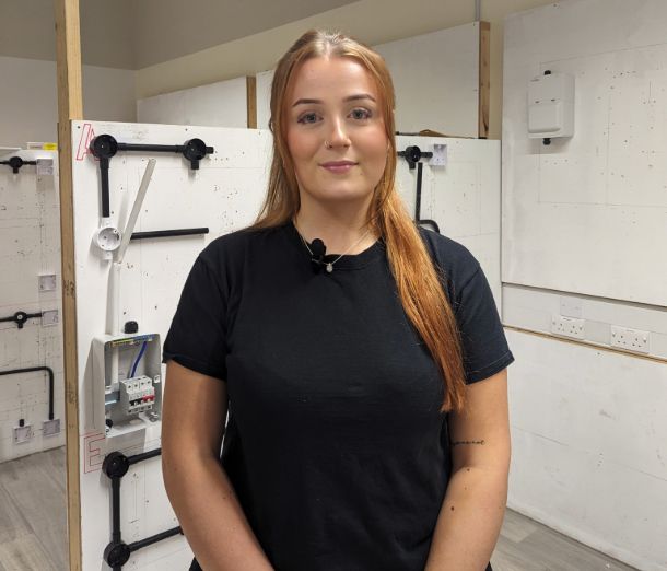 Electrician apprentice, Ruby who is studying at Leicester College, standing in a workshop at the college, wearing all black and smiling at the camera