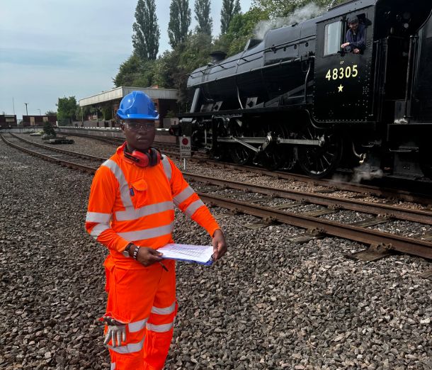 Leicester college student standing on the track bed next to a heritage steam train, wearing orange overalls and holding a clipboard