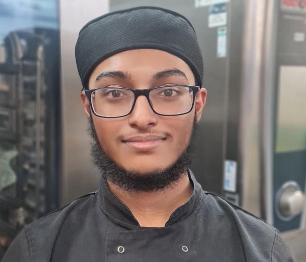 Leicester College student Shafiur, wearing black chef shirt, hat and glasses, smiling at the camera