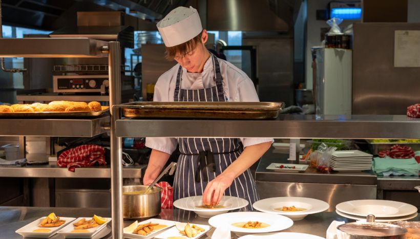 A catering student from Leicester College preparing meals in an industrial kitchen, wearing a chef hat and apron