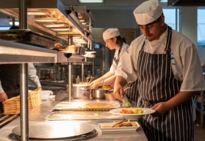 Catering students from Leicester College preparing meals in an industrial kitchen, wearing chef hats and aprons