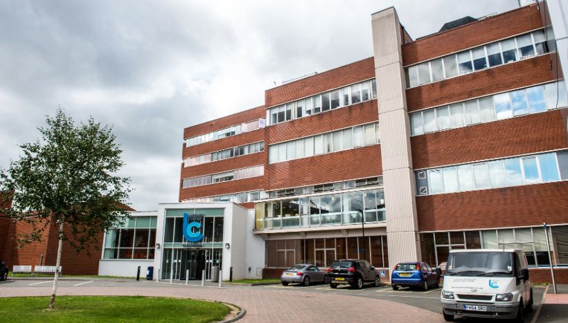 Image of the front of Leicester College's Freemens park campus - a large brick building with the college's logo on the front and vehicles parked outside