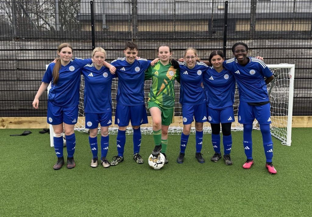 women in the LCFC blue football kit with goalkeeper in centre in green standing together in front of goal post