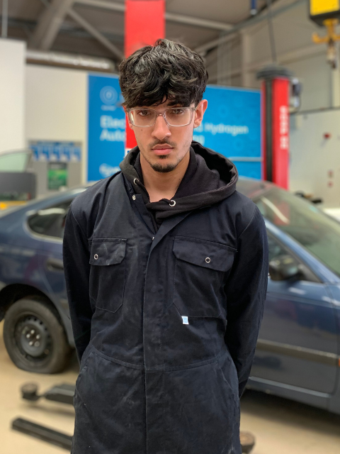 Student standing in the motor vehicle workshop in dark blue overalls